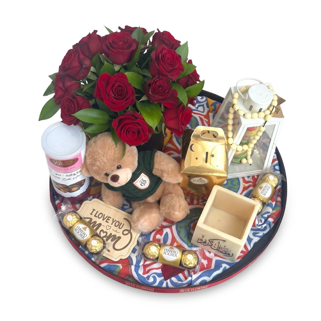 A round gift tray arrangement featuring a bouquet of red roses, a small teddy bear in a green sweater, assorted Ferrero Rocher chocolates, a gold pig bank, a decorative lantern with beads, and a card that reads 'I LOVE YOU MOM', all arranged on a patterned tray against a white background.